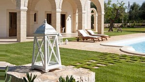A poolside area with lounge chairs, a white lantern, and a stone building with arches. - Dimora Donna Lucrezia (Fasano)