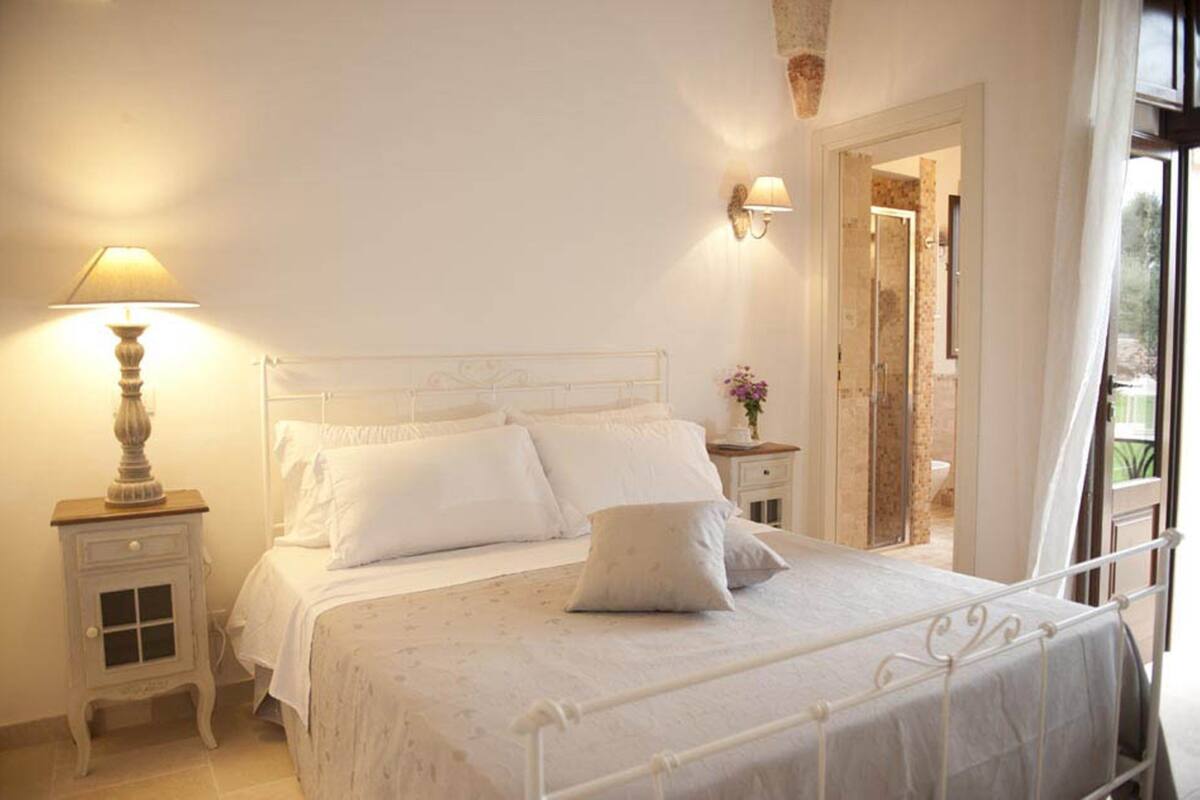 A neatly made bed with white linens and pillows, a bedside lamp, and a small bedside table with a lamp.