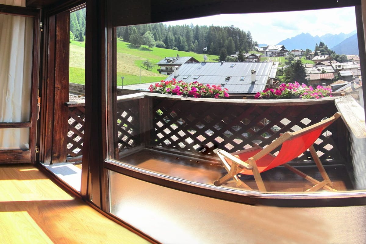 A bedroom with a bed, wooden walls, a window with a view of a village and mountains, and a balcony with flowers.