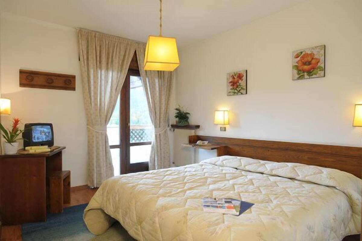 A hotel room with a large bed, a desk with a television, a window with curtains, and wall-mounted lamps.