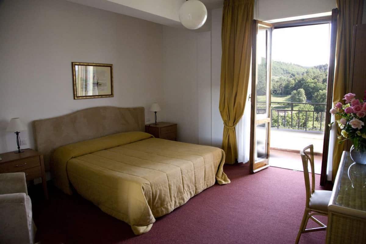 A hotel room with a bed, a nightstand, a chair, a table with a vase of flowers, and a balcony with a view of greenery.