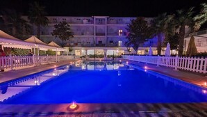 Una piscina nocturna con valla blanca, palmeras y un edificio al fondo.