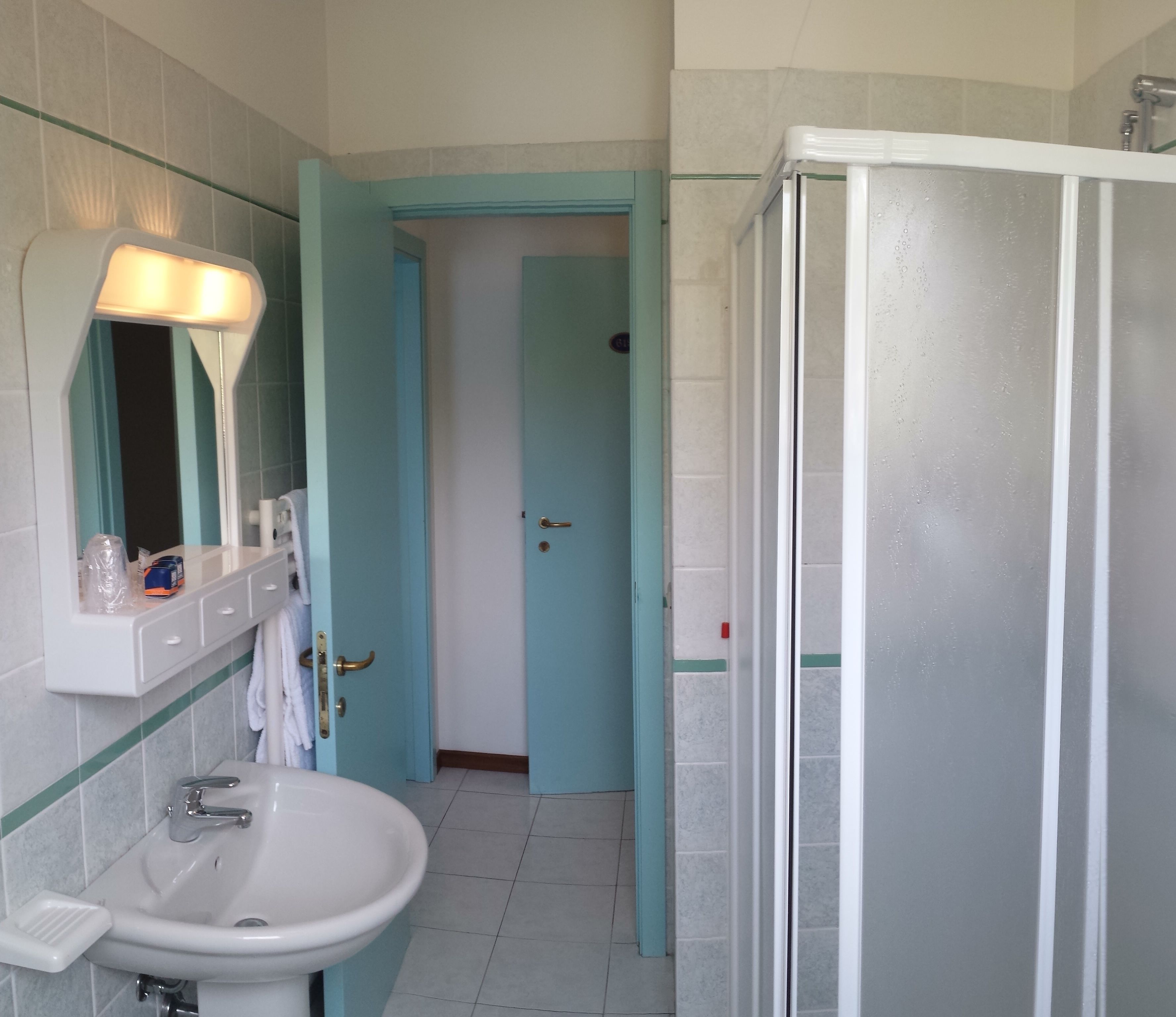 Un baño con un lavamanos blanco, un espejo, una lámpara, cabina de ducha y una puerta que da a otra habitación.