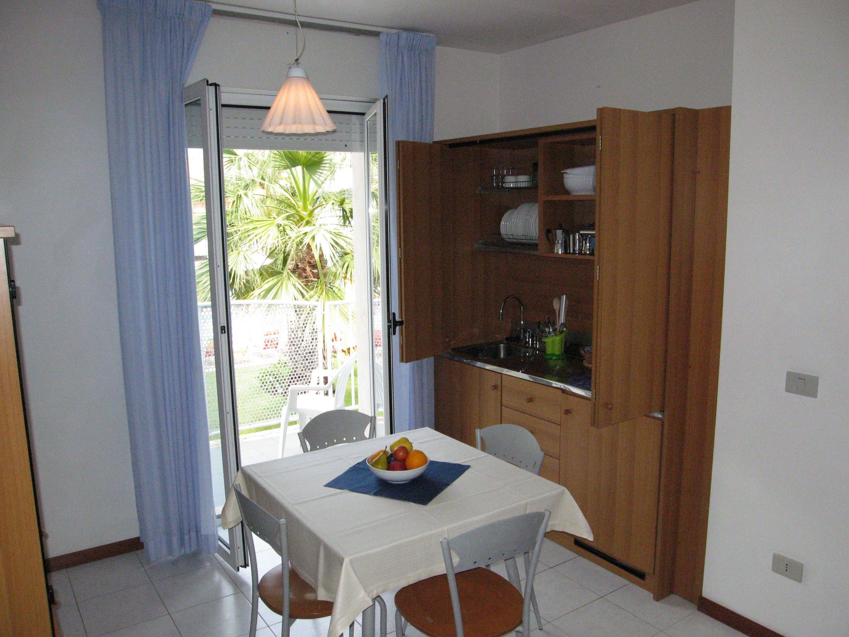 Une cuisine compacte avec un coin repas, comprenant une table avec des chaises, un panier de fruits et une vue sur un jardin par une porte ouverte.