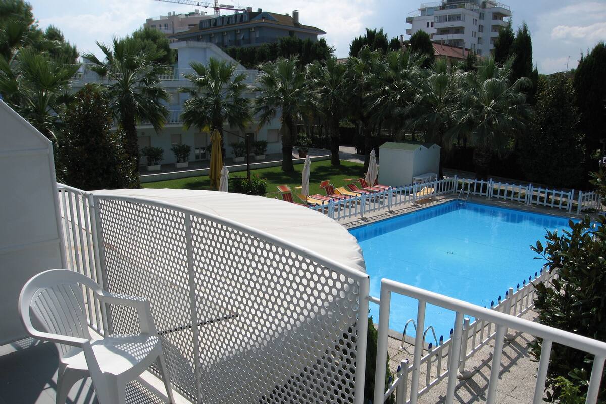 A swimming pool with a white fence, surrounded by palm trees and a white building.