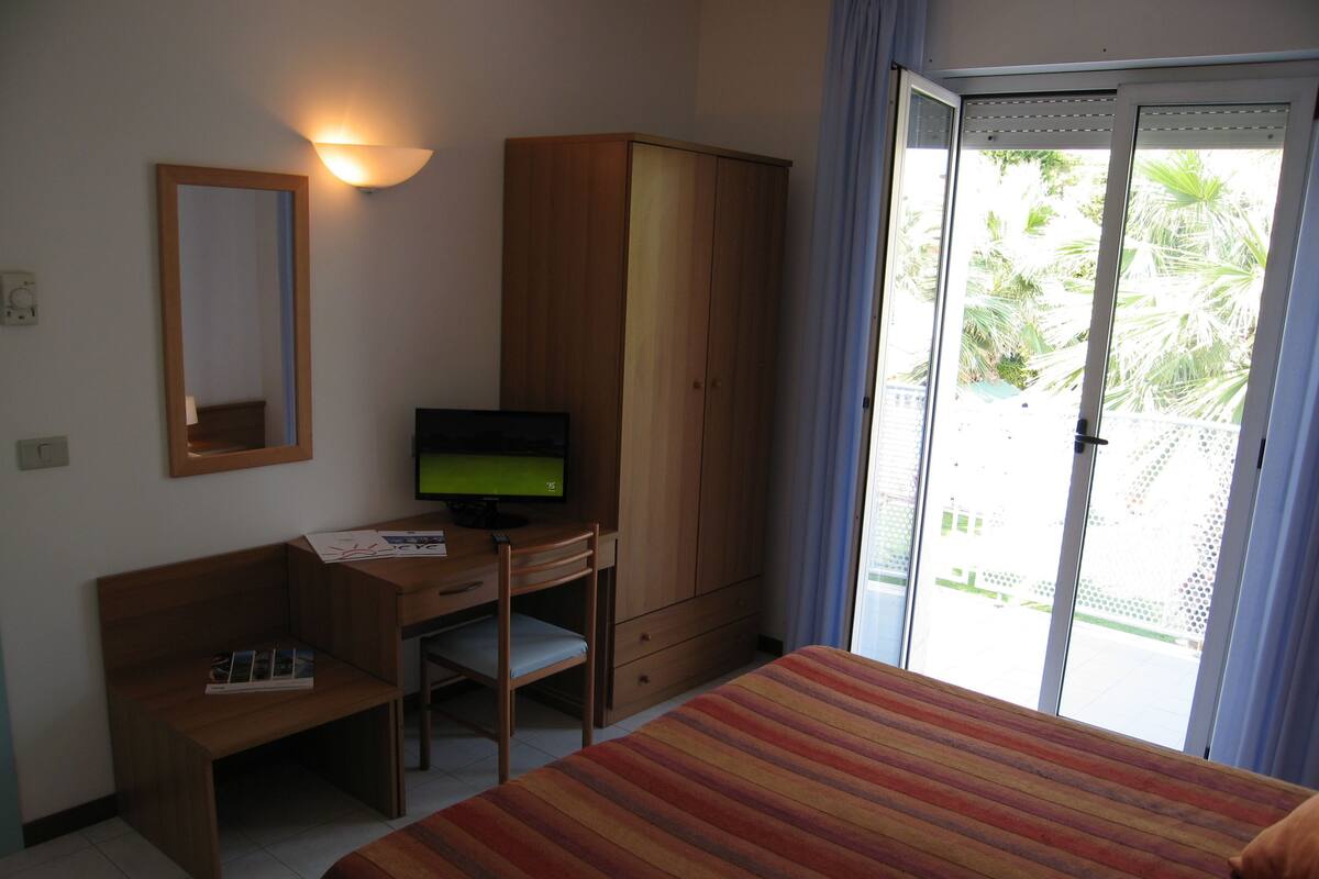 A hotel room with a bed, a desk with a computer, a wardrobe, and a balcony with a view of greenery.