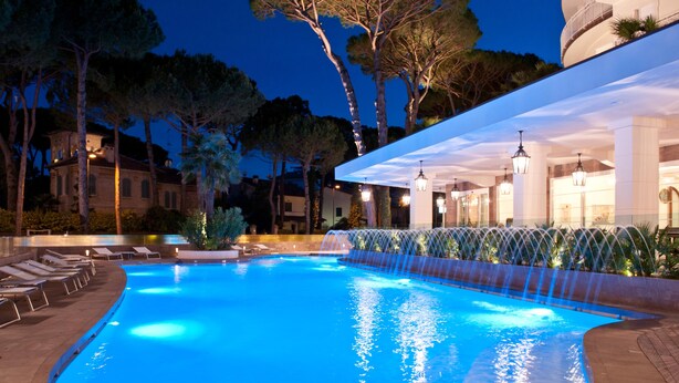 A swimming pool with a waterfall feature, lounge chairs, and a covered area with columns and lighting. - Hotel Belvedere (Cervia)