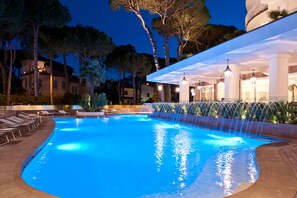 A swimming pool with a waterfall feature, lounge chairs, and a covered area with columns and lighting. - Hotel Belvedere (Cervia)