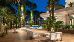 An outdoor pool area with wooden decking, white lounge chairs, and a pool in the background. - Hotel Belvedere (Cervia)