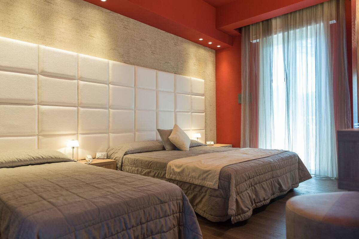 A hotel room with two beds, a large padded headboard, a nightstand with a lamp, and a window with sheer curtains.