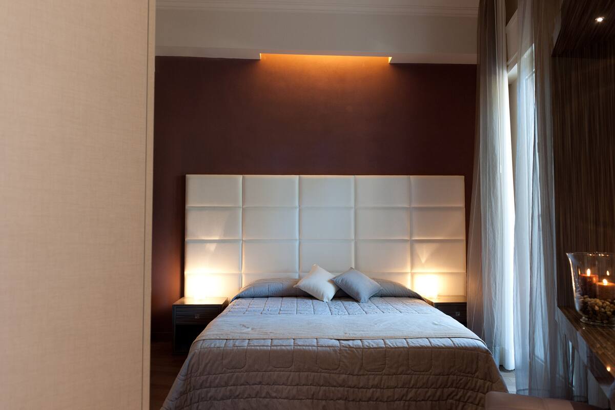 A bedroom with a large padded headboard, a bed with a quilted cover, and a bedside table with a lamp.