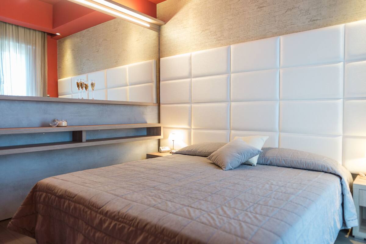 A modern bedroom with a large bed, a bedside table, a nightstand, and a wall-mounted shelf with decorative items.