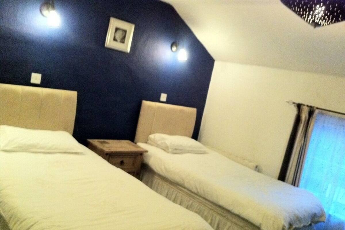Quarto de hotel com duas camas de solteiro, uma pequena mesa de cabeceira e uma janela com cortinas.