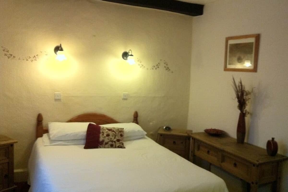 Cama bem arrumada com cabeceira de madeira, duas luminárias fixadas na parede, uma mesinha de cabeceira e um criado-mudo.