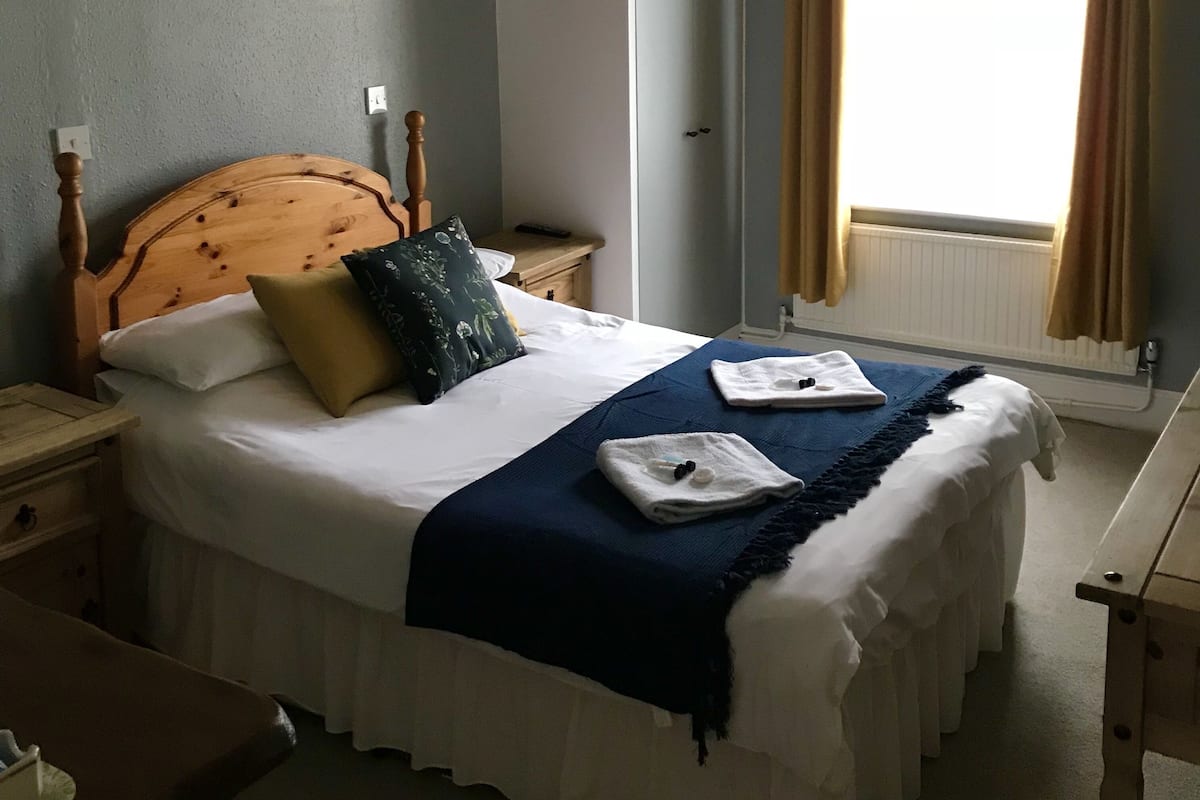 Cama bem arrumada com cabeceira de madeira, roupa de cama branca, manta azul e duas toalhas dobradas.