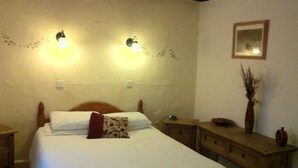 A neatly made bed with white bedding and a red pillow, two wall-mounted lights, a wooden nightstand, a small table with a vase, and a framed picture on the wall.