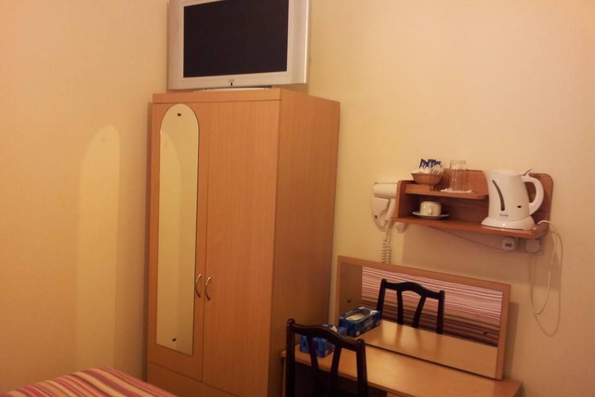 A bedroom with a bed, a wooden wardrobe, a desk with a chair, a television on top of the wardrobe, and a shelf with a kettle and cups.