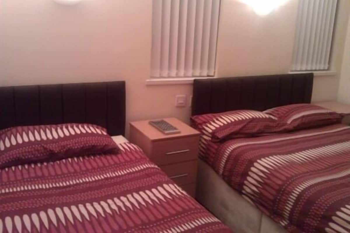 A bedroom with two beds, each with a patterned bedspread, a nightstand with a remote control, and a window with blinds.