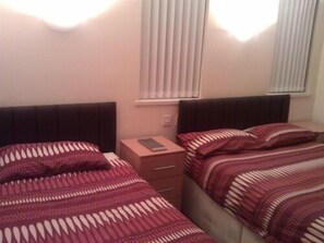 A bedroom with two beds, each with a patterned bedspread, a nightstand with a remote control, and a window with blinds.