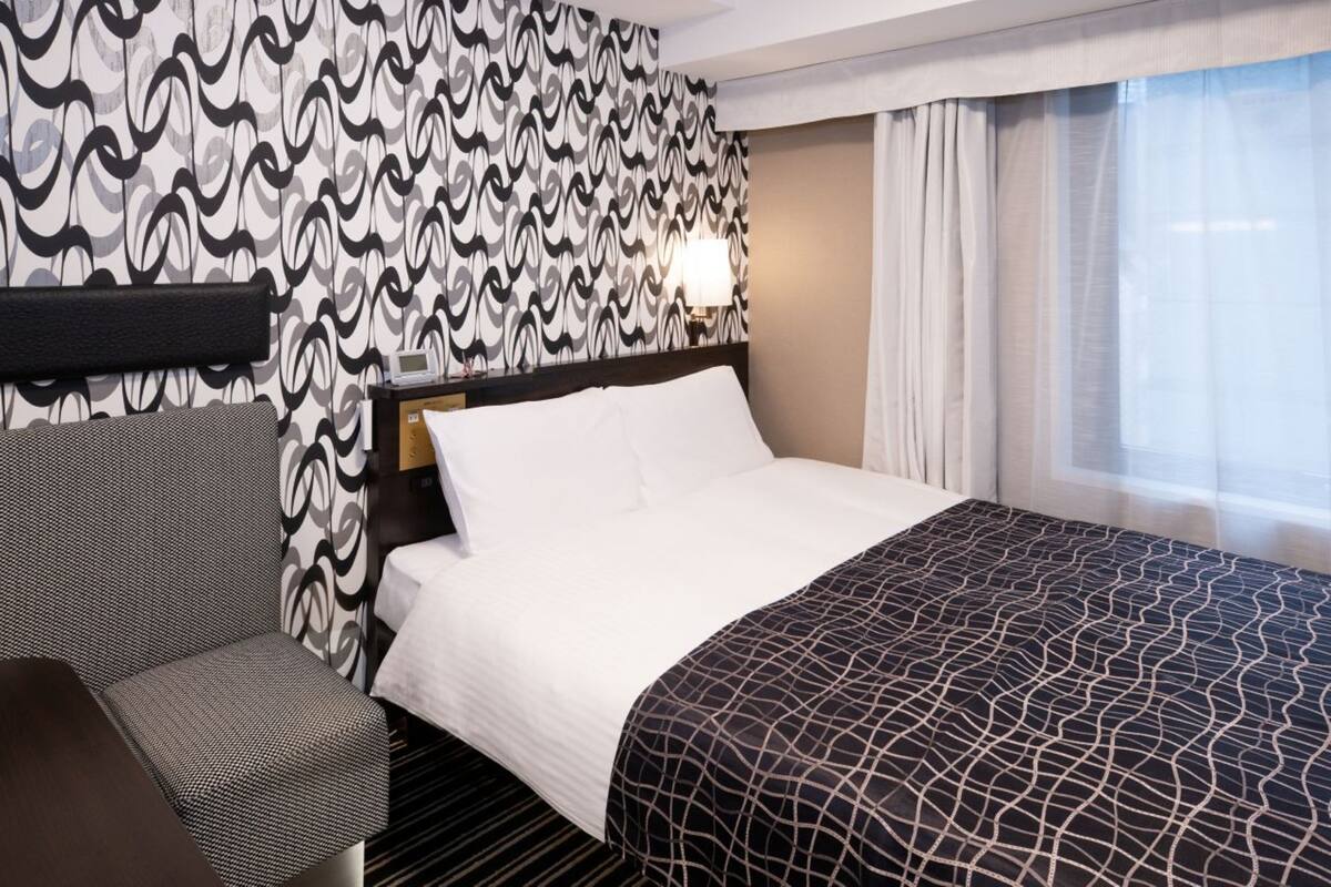 A hotel room with a patterned wallpaper, a bed with a patterned bedspread, a bedside table, a chair, and a window with curtains.