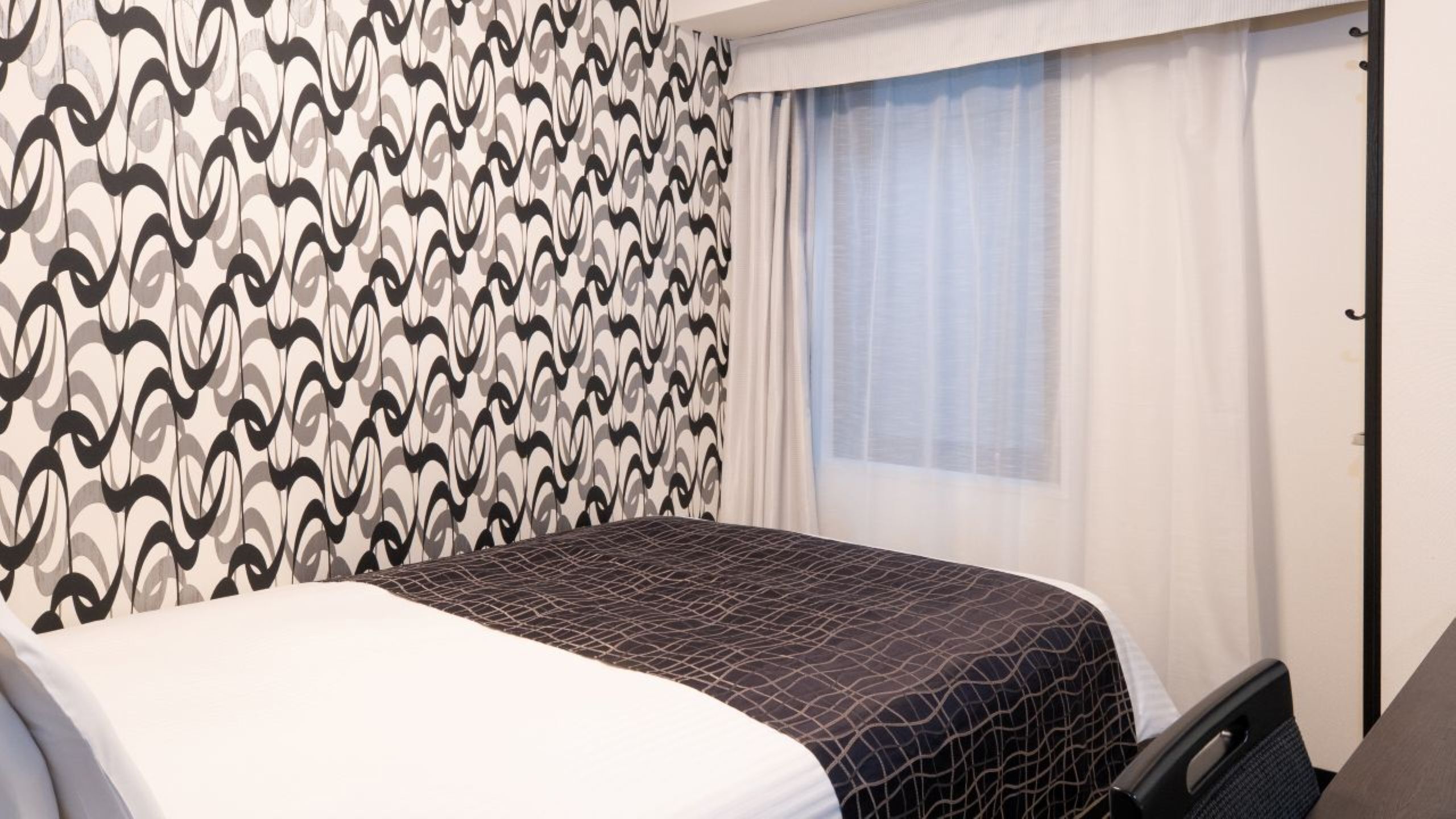 Quarto de hotel com parede estampada, cama com colcha de padrão geométrico, janela com cortinas translúcidas e uma cadeira.