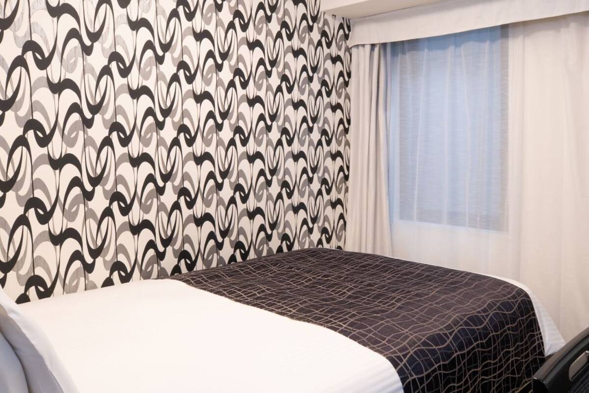 A hotel room with a patterned wall, a bed with a geometric-patterned bedspread, a window with sheer curtains, and a chair.