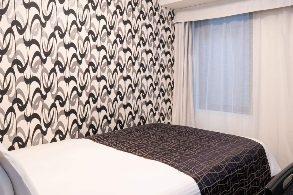 Ett hotellrum med en vägg som har ett mönster, en säng med ett geometriskt mönstrat överkast, ett fönster med tunna, genomskinliga gardiner och en stol.