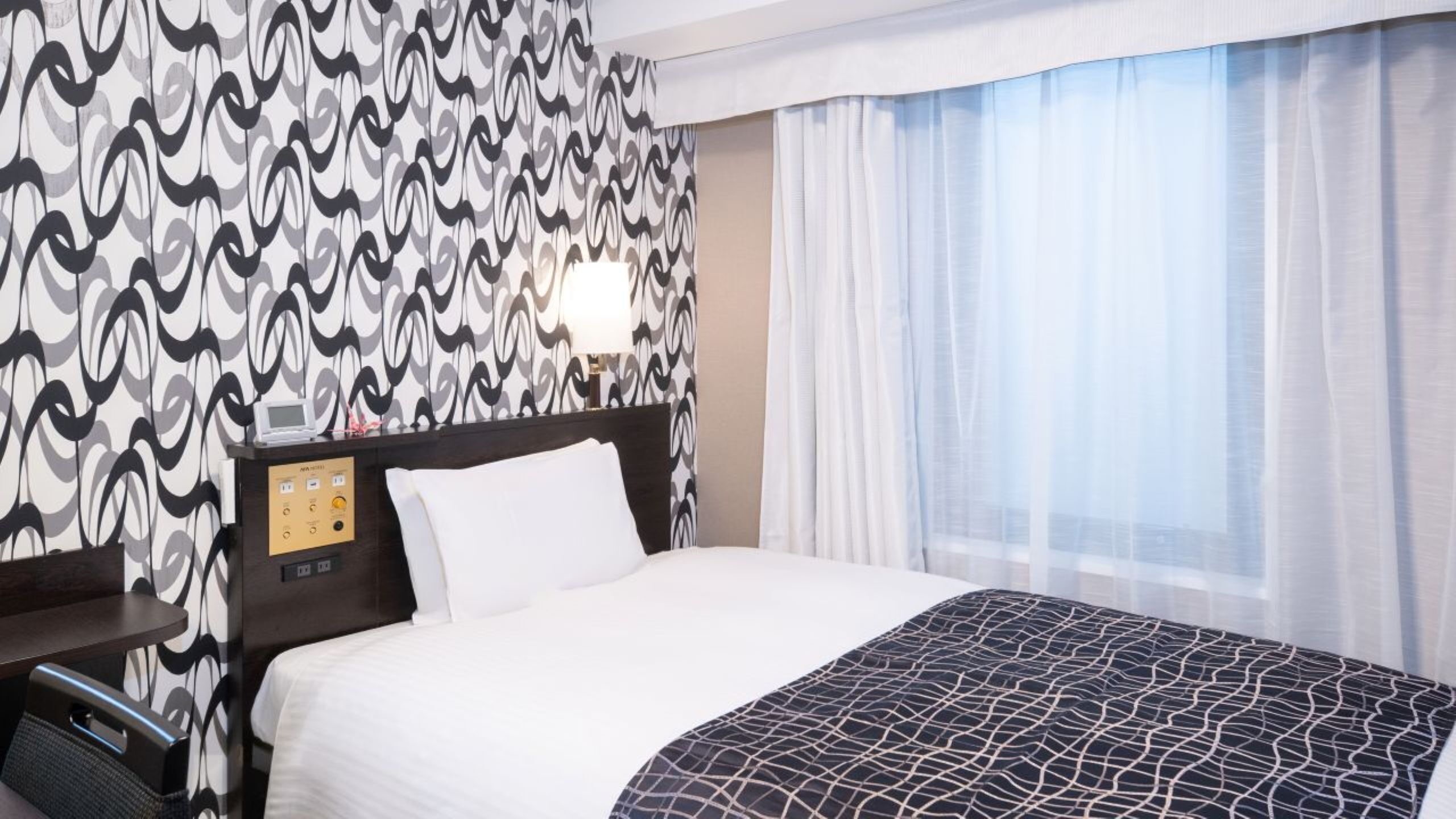 Quarto de hotel com papel de parede estampado, cama com colcha de padrão preto e branco, mesa de cabeceira com abajur e janela com cortinas leves.