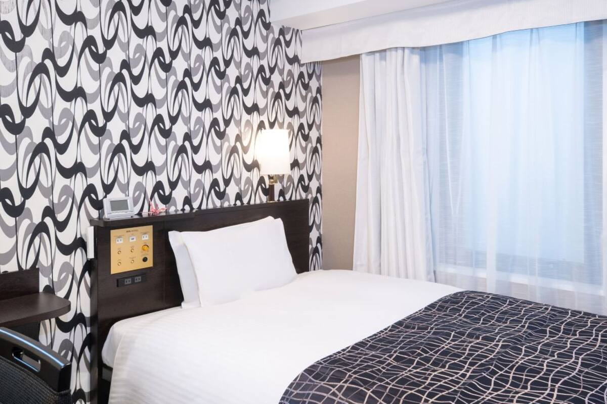 A hotel room with a patterned wallpaper, a bed with a black and white patterned bedspread, a bedside table with a lamp, and a window with sheer curtains.