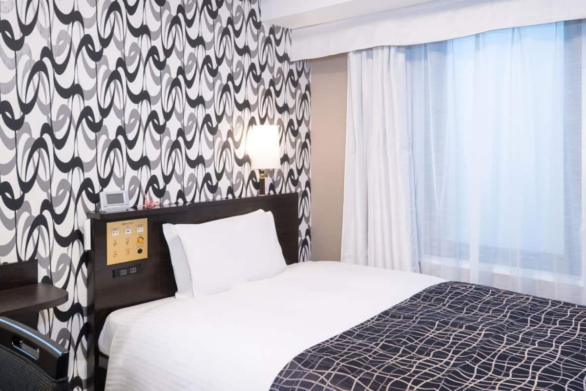 Ett hotellrum med tapet med ett mönster, en säng med ett svartvitt mönstrat överkast, ett sängbord med en lampa och ett fönster med tunna, genomskinliga gardiner.