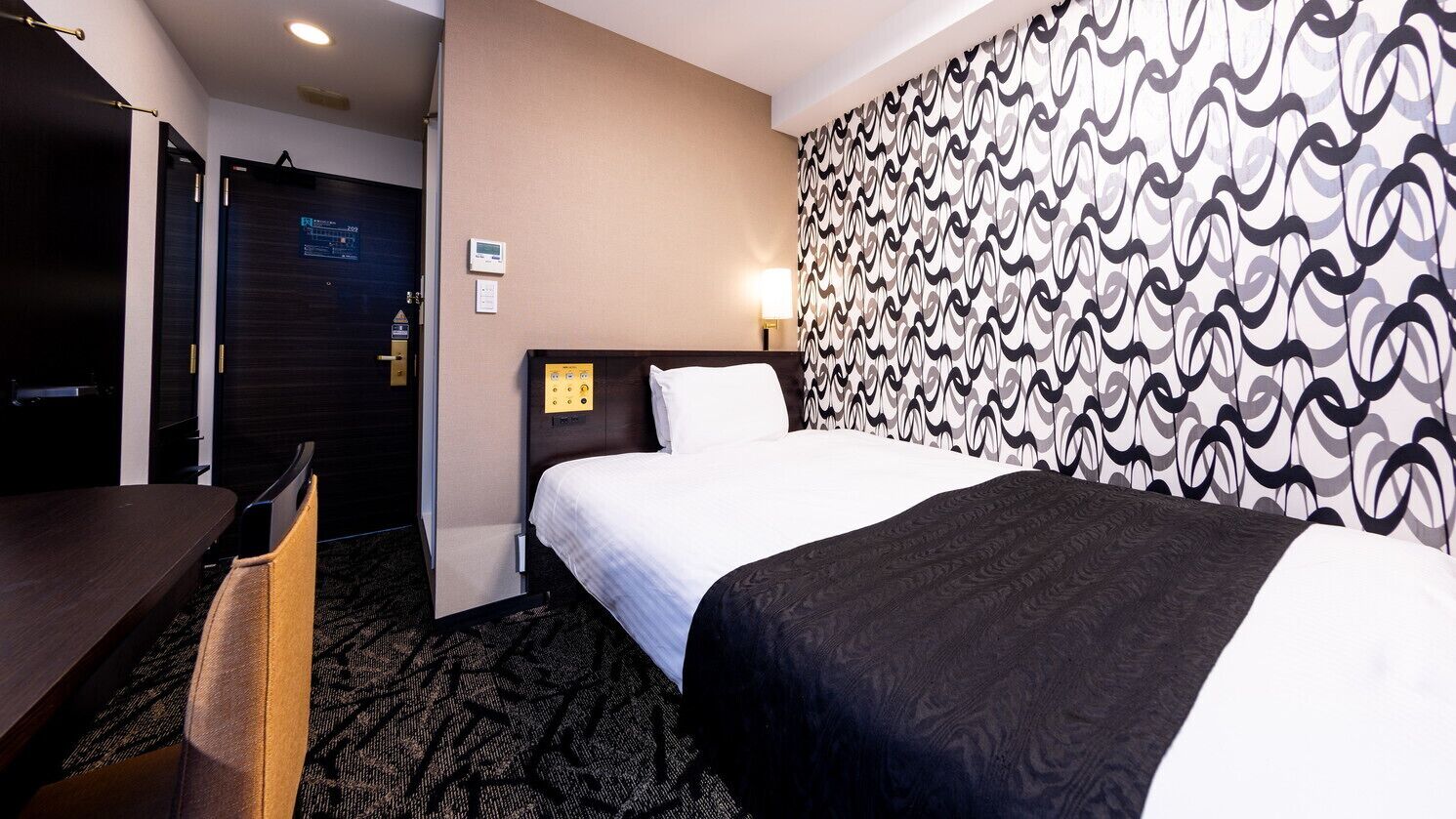 A hotel room with a bed, a desk, a chair, a patterned wall, and a door with a sign.