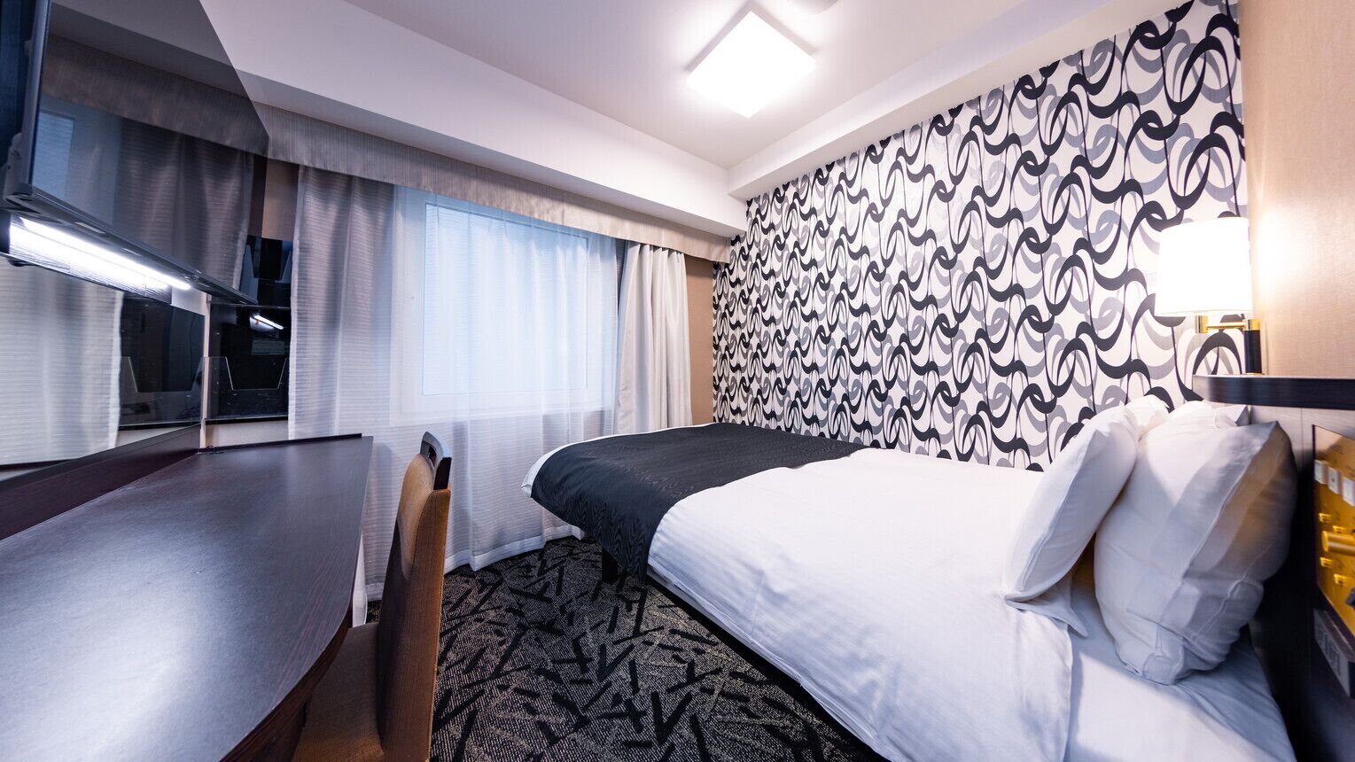 A hotel room with a bed, a desk, a chair, a patterned wall, and a window with curtains.