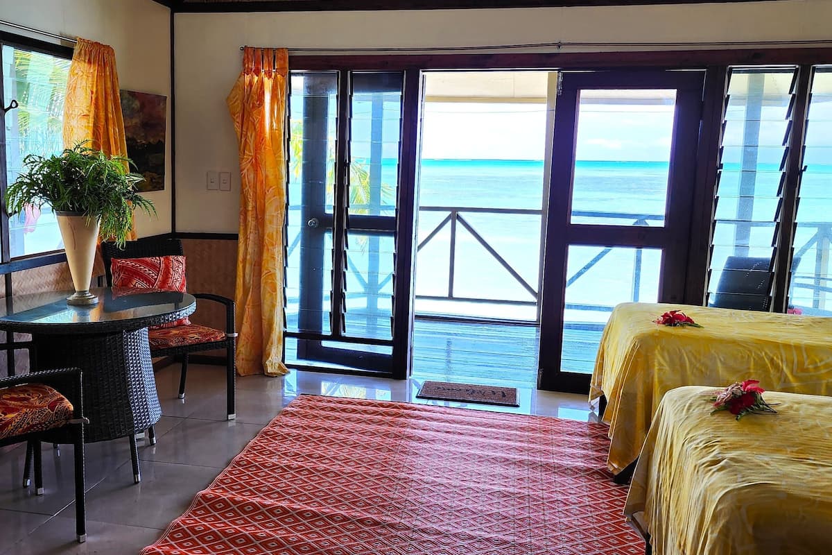 A room with a bed, a table with chairs, a potted plant, and a view of the ocean through large windows.