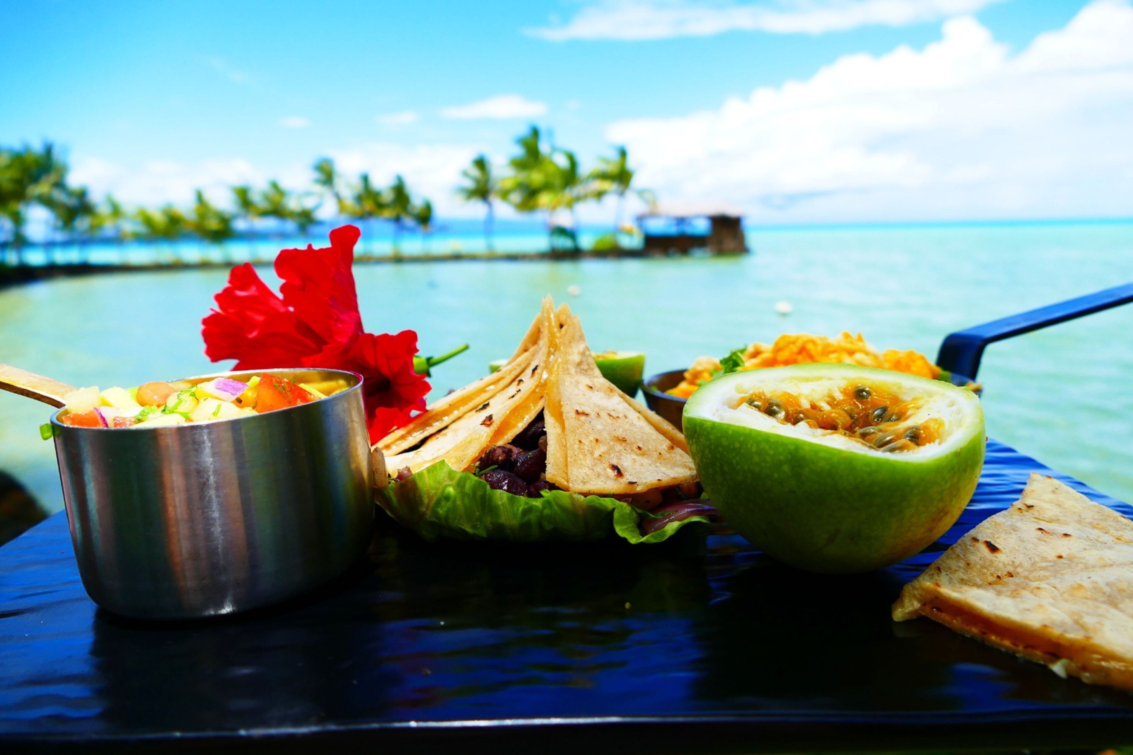 Comida en un entorno tropical, incluyendo un tazón de ensalada, un vegetal relleno y media fruta en un plato azul.
