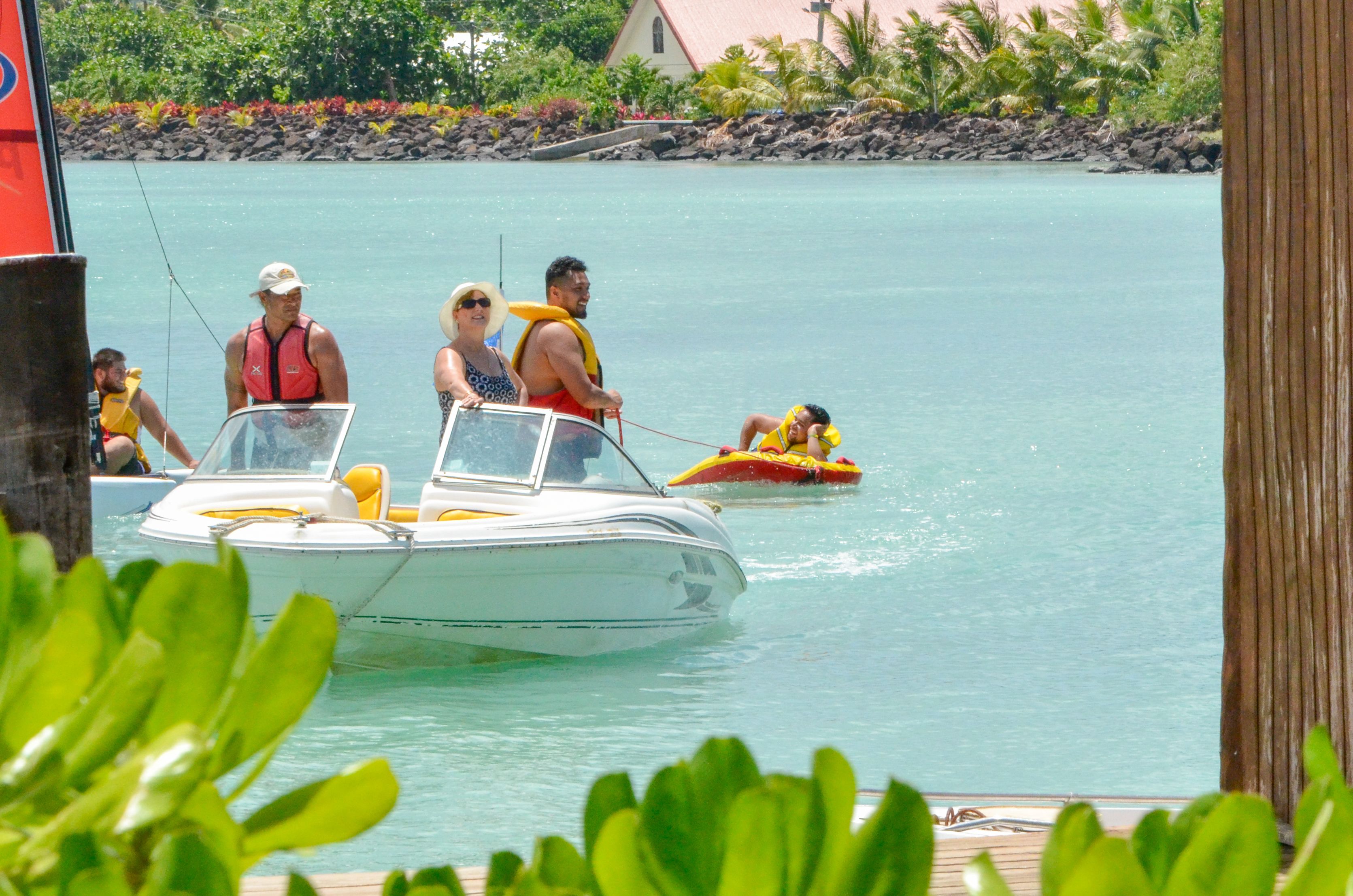 Un bote con pasajeros usando chalecos salvavidas, un pequeño bote inflable y un muelle con follaje al fondo.