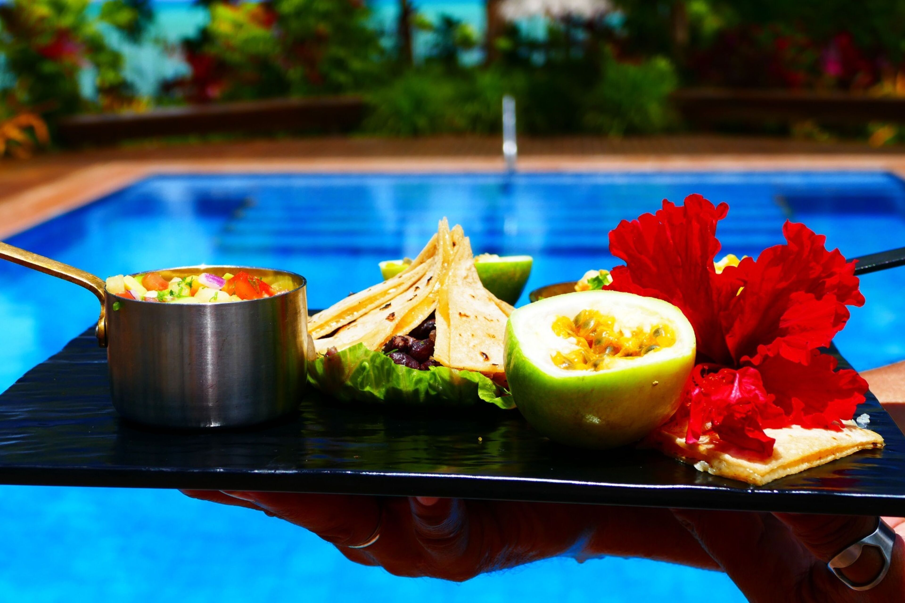 A dining table with a salad, tortilla chips, and a halved fruit next to a pool.