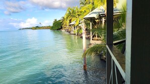 A tropical beach view with clear turquoise water, palm trees, and a wooden structure with a roof. - Le Vasa Resort (Cape Fatuosofia)