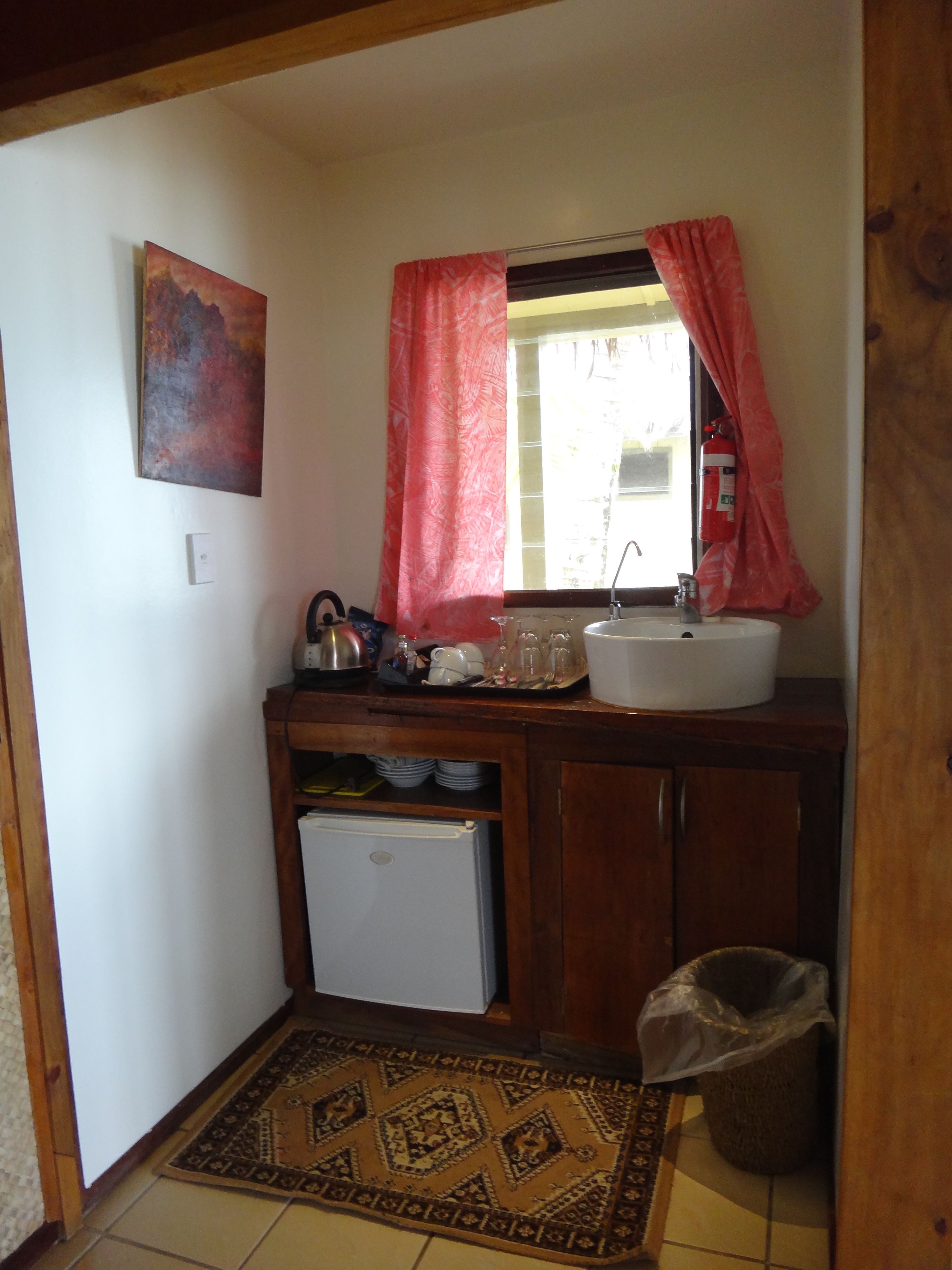 A compact kitchen with a white sink, a small refrigerator, and a window with red curtains.