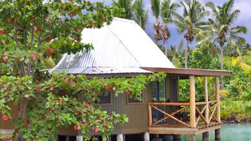 A wooden house with a metal roof, situated on stilts over water, surrounded by lush greenery and palm trees.