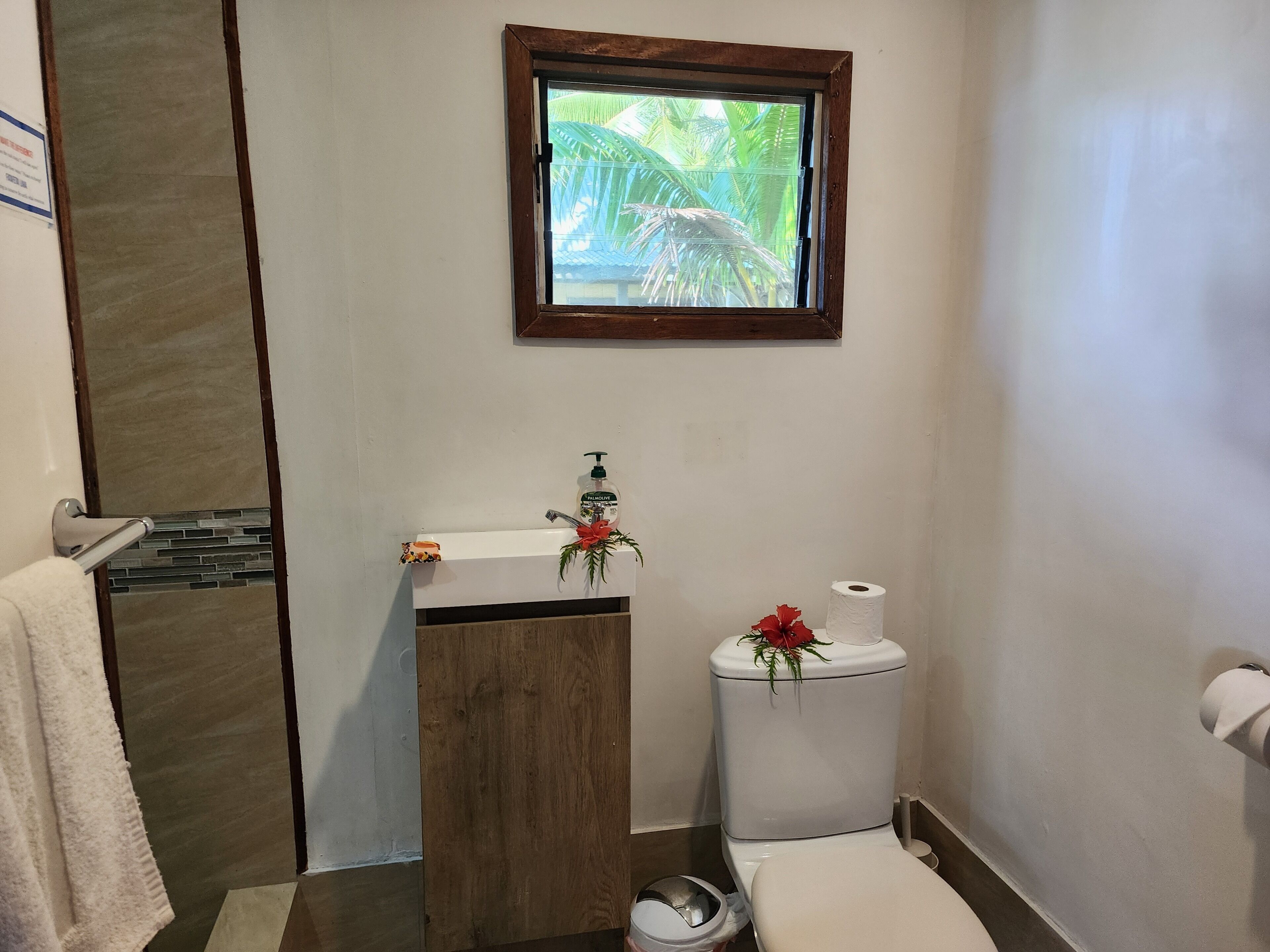 A bathroom with a wooden vanity, a toilet, and a window with a view of palm trees.