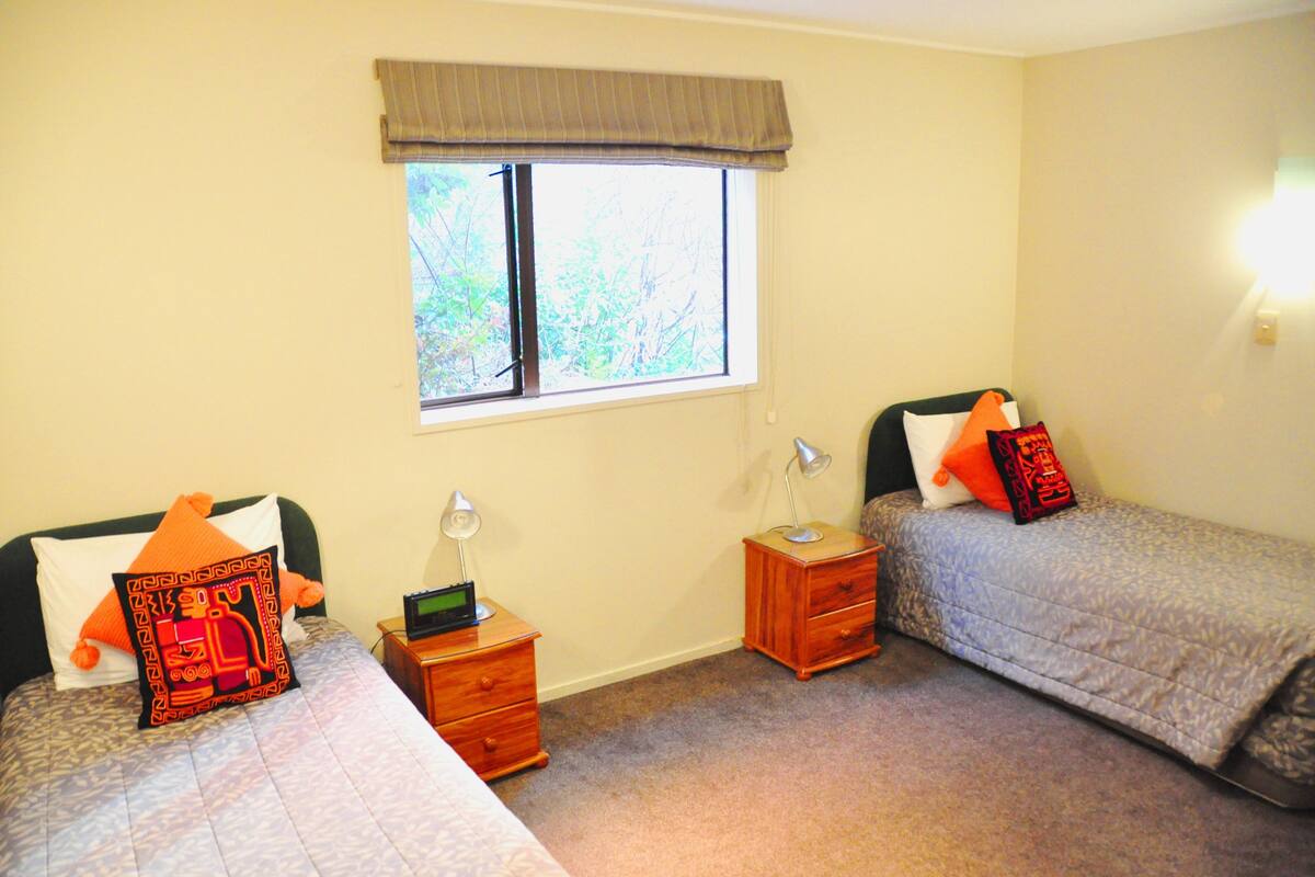 A bedroom with two single beds, a window with a view of trees, a bedside table with a lamp, and a nightstand with a clock.