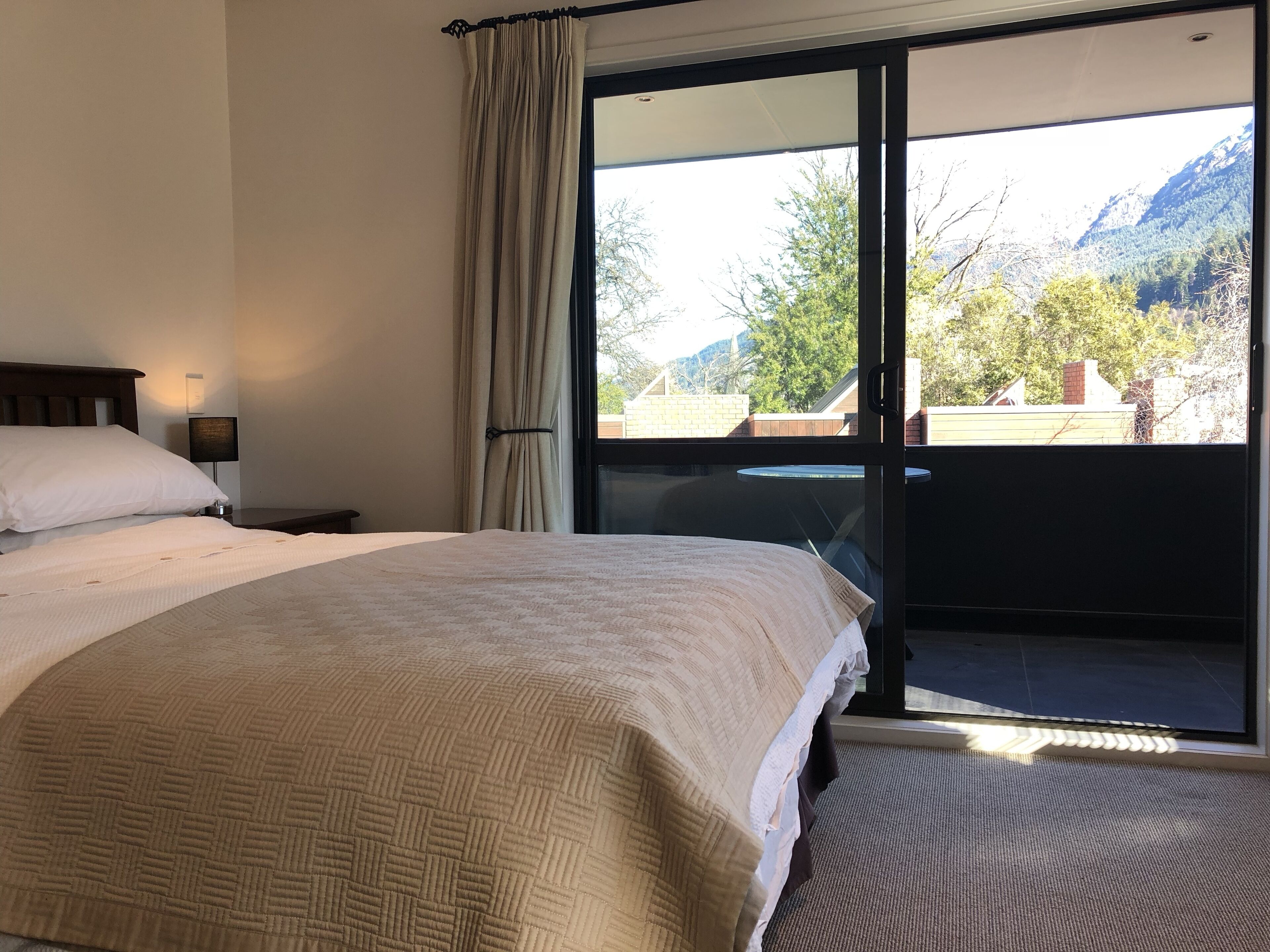 A bedroom with a large bed, a nightstand, a lamp, and a view of a balcony with mountains in the background.