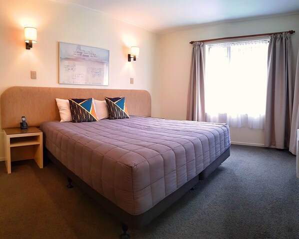 A neatly arranged hotel room with a large bed, a bedside table with a phone, two wall-mounted lamps, a framed picture on the wall, and a window with curtains.