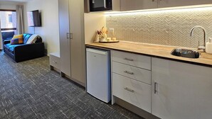 Cocina moderna con microondas, fregadero y lavavajillas. Hay un comedor con sofá y televisor. El piso está alfombrado y la salpicadura trasera es de baldosas en espiga.