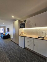A modern kitchen with a microwave, sink, and dishwasher. There is a dining area with a sofa and a TV. The room has a carpeted floor and a herringbone tile backsplash.