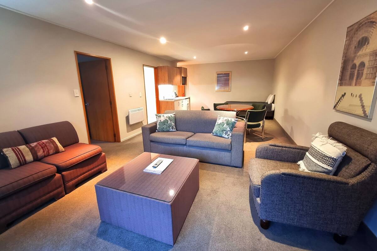 A modern living room with a sofa, armchair, and ottoman. A dining table and chairs are visible in the background.