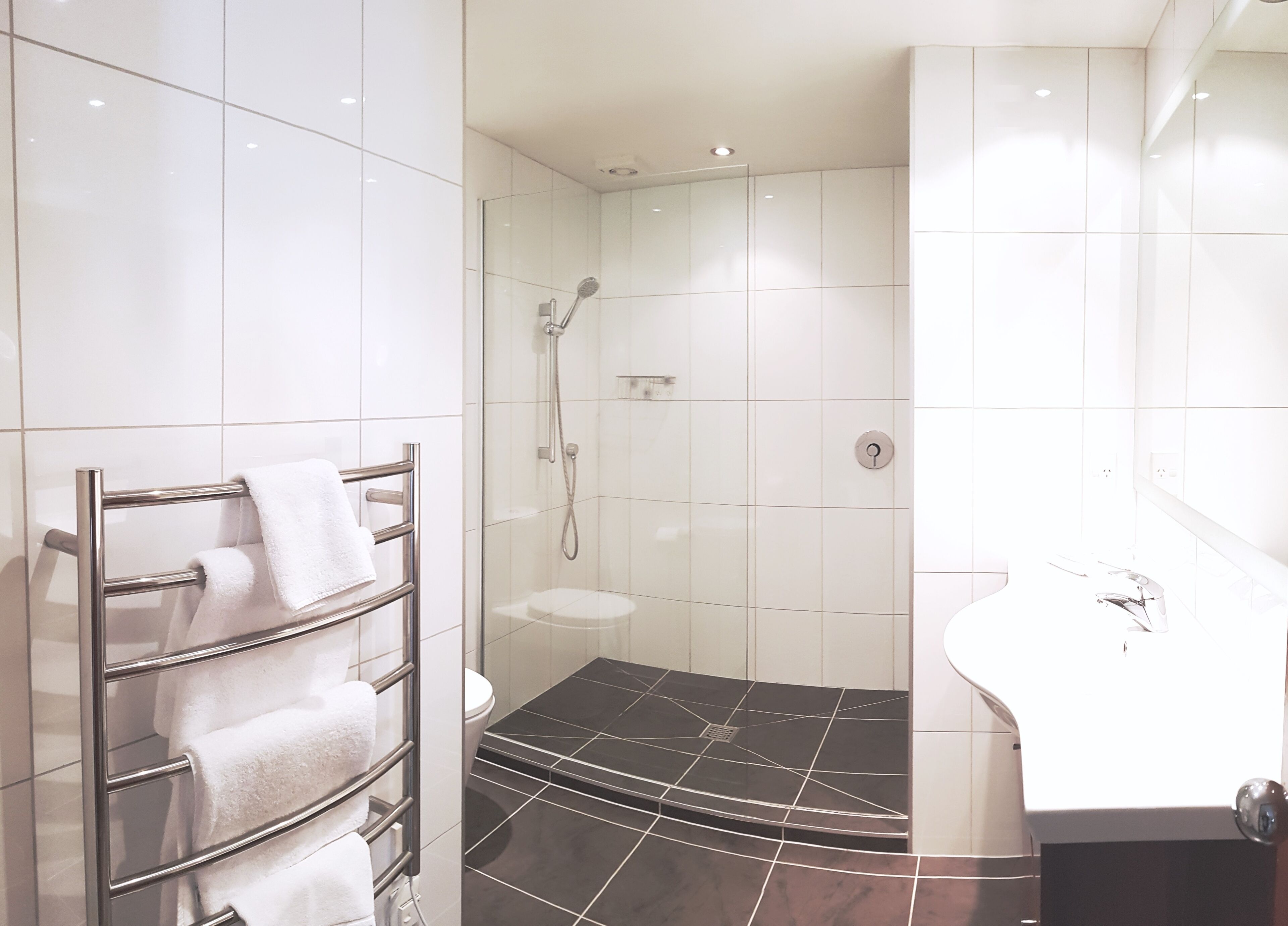 Un baño moderno con ducha envuelta en vidrio, un lavabo blanco y un perchero con varias toallas.