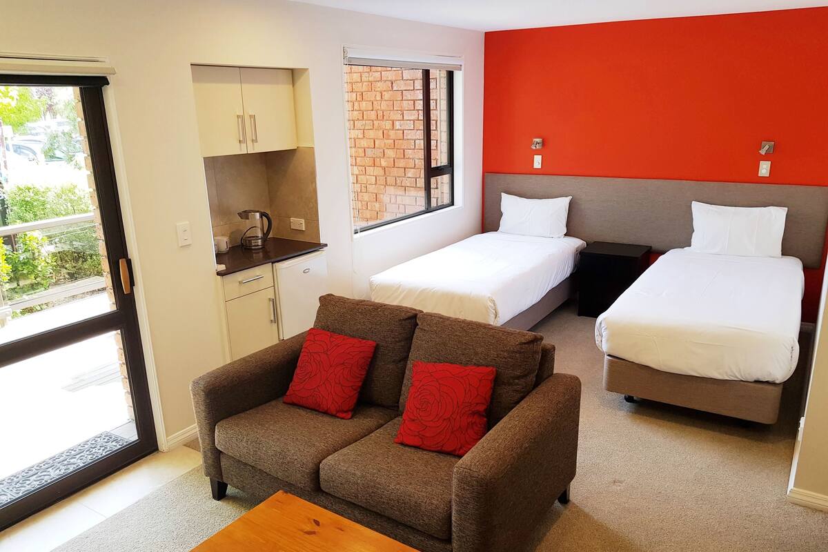 A compact room with a kitchenette, two single beds, a sofa with red cushions, and a wooden table.