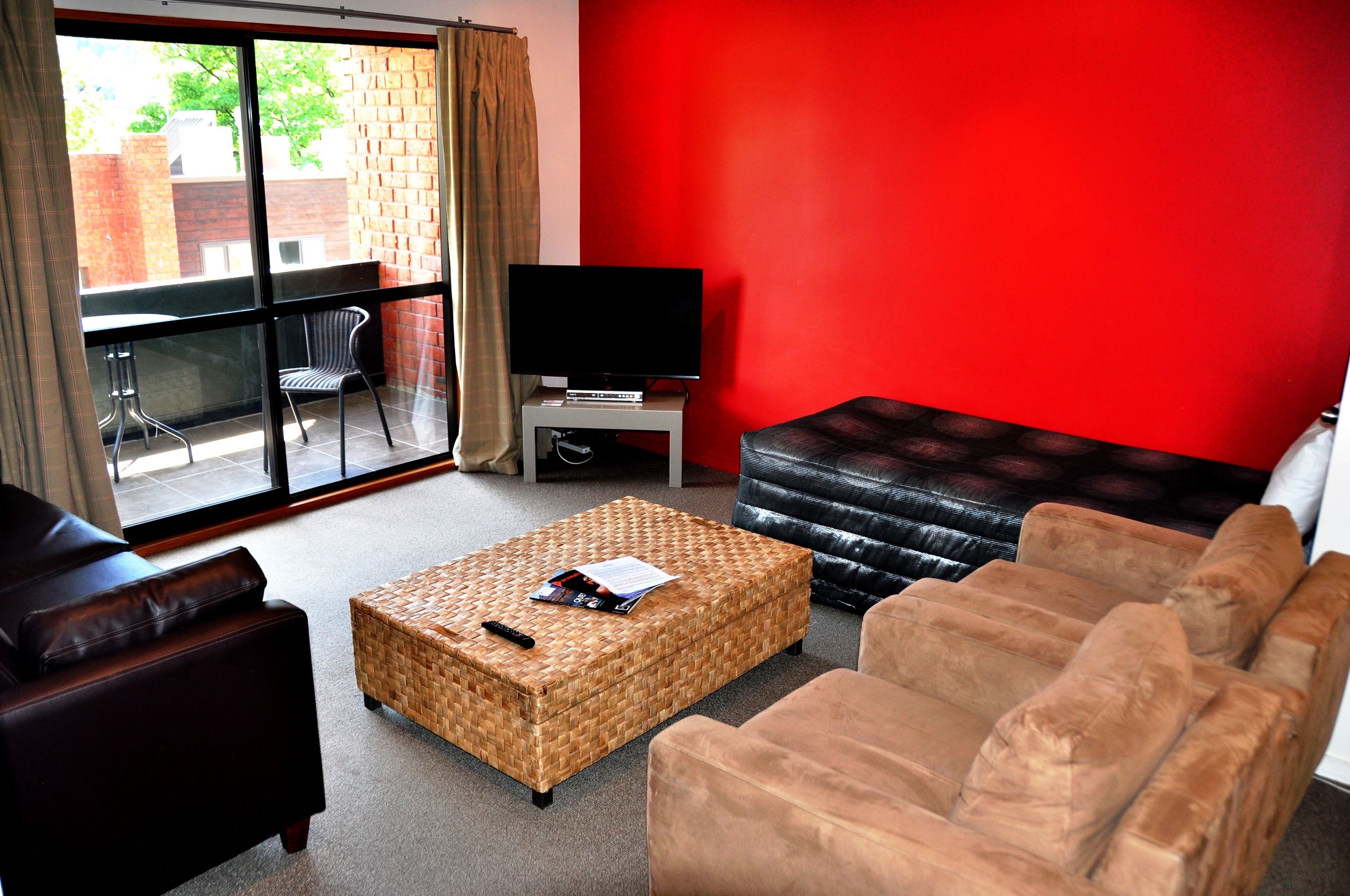 A living room with a red wall, a television on a stand, a wicker ottoman, a black leather sofa, and a brown leather couch.