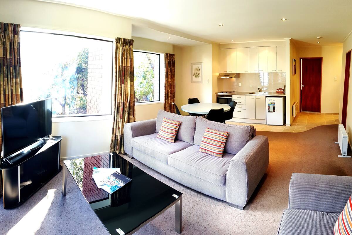 A modern living room with a sofa, a coffee table, and a TV. There is a kitchen area in the background with cabinets and a refrigerator.